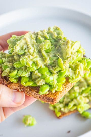 Hand holding a piece of toast topped with edamame and avocado.